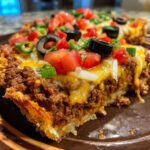 A close-up slice of 2-layer mexican pizza showing seasoned ground beef, melted cheese, tomatoes, olives, and green onions.