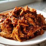 A close-up of saucy, shredded pulled chicken piled high on a white plate, ready to eat.