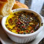 A close-up of a white ramekin filled with vibrant olive oil bread dip, seasoned heavily with herbs and red pepper flakes, next to torn bread.