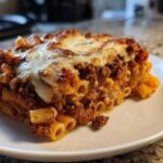A close-up of a hearty slice of meat pasta bake with melted, browned cheese on top, served on a white plate.