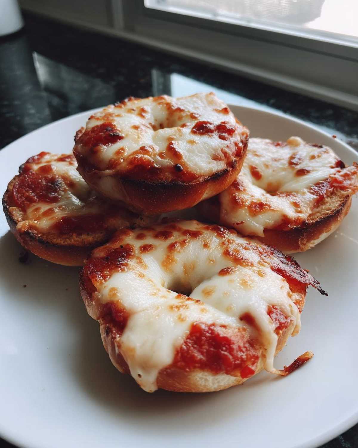 Four freshly made pizza bagels topped with melted mozzarella cheese and sauce, served on a white plate.