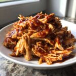 A close-up of tender, shredded pulled chicken generously coated in rich BBQ sauce, served in a white bowl.