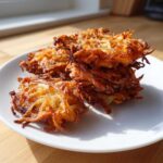 A stack of crispy, golden brown Amish Onion Fritters piled on a white plate in bright sunlight.