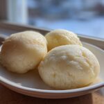 Three perfectly shaped scoops of pale yellow fufu served on a small white dish near a window.