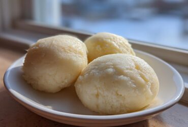 Three perfectly shaped scoops of pale yellow fufu served on a small white dish near a window.