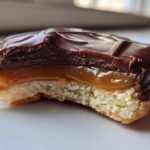 A close-up view of a homemade Twix cookie with a bite taken out, showing the shortbread base, gooey caramel layer, and thick chocolate topping.
