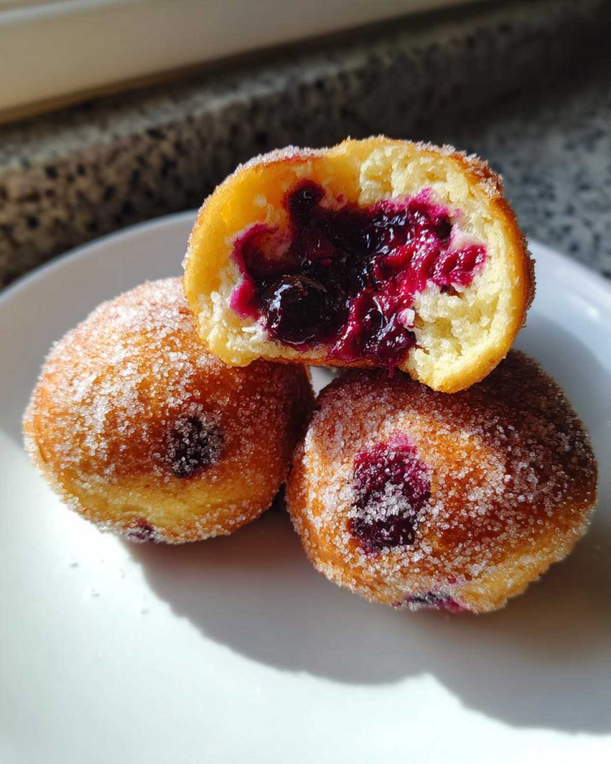 Three golden, sugar-coated Blueberry Pie Bombs on a white plate, one cut open revealing rich blueberry filling.