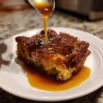A close-up of a slice of bread pudding with bourbon sauce being drizzled from a spoon.