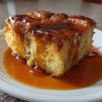 A square slice of rich bread pudding with bourbon sauce generously drizzled over the top and pooled on the white plate.