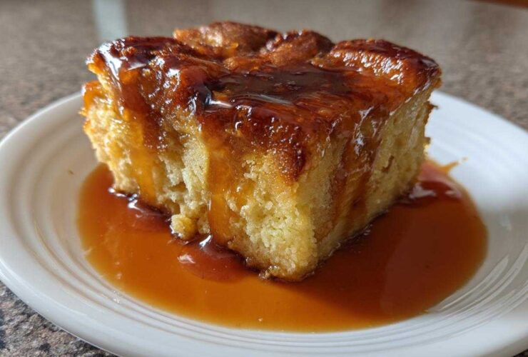 A square slice of rich bread pudding with bourbon sauce generously drizzled over the top and pooled on the white plate.