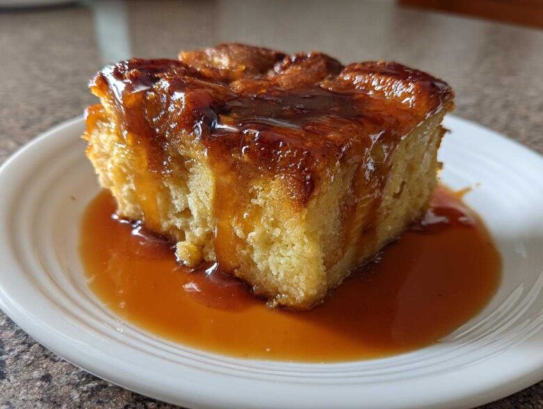A square slice of rich bread pudding with bourbon sauce generously drizzled over the top and pooled on the white plate.