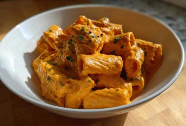 A white bowl filled with rigatoni pasta coated in vibrant orange butternut squash pasta sauce, garnished with herbs.