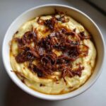A white bowl filled with creamy caramelized onion dip, generously topped with dark, crispy fried onions.
