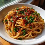 Close-up of a white bowl filled with spaghetti tossed in sauce and topped generously with caramelized onion pasta and fresh parsley.