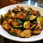 A close-up of a white bowl filled with glossy chicken zucchini stir fry coated in a savory sauce.