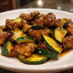 Close-up of glossy, saucy chicken zucchini stir fry pieces garnished with sesame seeds on a white plate.