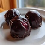 Close-up of three glossy, dark chocolate covered cherries on a white plate, one showing the bright red cherry inside.