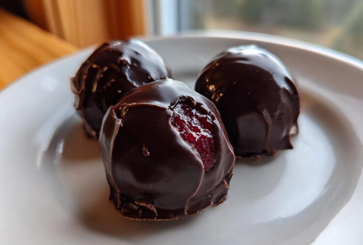 Close-up of three glossy, dark chocolate covered cherries on a white plate, one showing the bright red cherry inside.