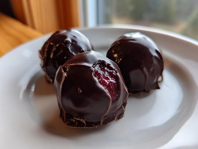 Close-up of three glossy, dark chocolate covered cherries on a white plate, one showing the bright red cherry inside.