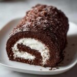 A cross-section view of a decadent chocolate yule log cake filled with white cream and covered in chocolate shavings.