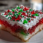 Close-up of a slice of Christmas poke cake featuring layers of white cake, red and green gelatin, whipped topping, and holiday sprinkles.