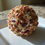 A close-up of a perfectly formed cranberry pecan cheese ball coated in chopped nuts and dried cranberries.