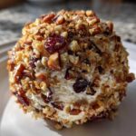 A close-up view of a perfectly formed cranberry pecan cheese ball coated in chopped nuts and dried cranberries.