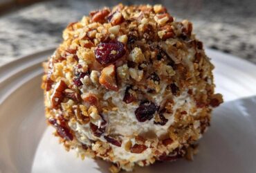 A close-up view of a perfectly formed cranberry pecan cheese ball coated in chopped nuts and dried cranberries.