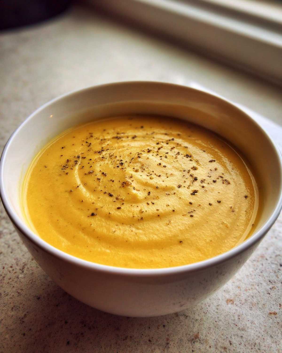 A close-up of a bowl of creamy, yellow cauliflower soup topped with freshly cracked black pepper.