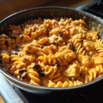 Close-up of creamy enchilada pasta with rotini noodles, chicken, and black beans in a dark skillet.