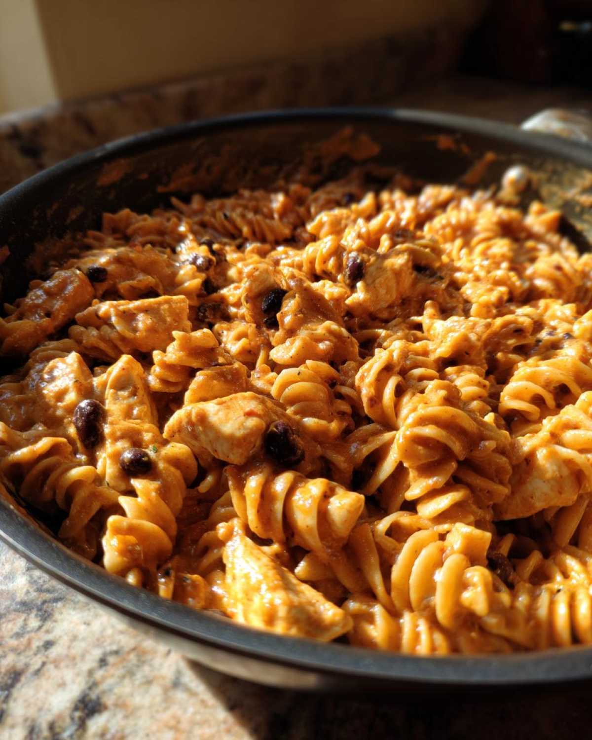 Close-up of creamy enchilada pasta with rotini noodles, chicken chunks, and black beans in a dark skillet.