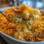 A spoonful of creamy funeral potatoes recipe lifted from a white casserole dish, showing a golden cornflake topping.