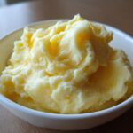 A close-up of a white bowl filled with rich, creamy, and fluffy mashed potatoes with melted butter visible on top.