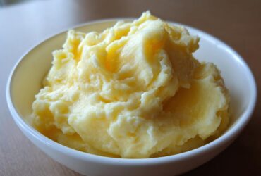 A close-up of a white bowl filled with rich, creamy, and fluffy mashed potatoes with melted butter visible on top.