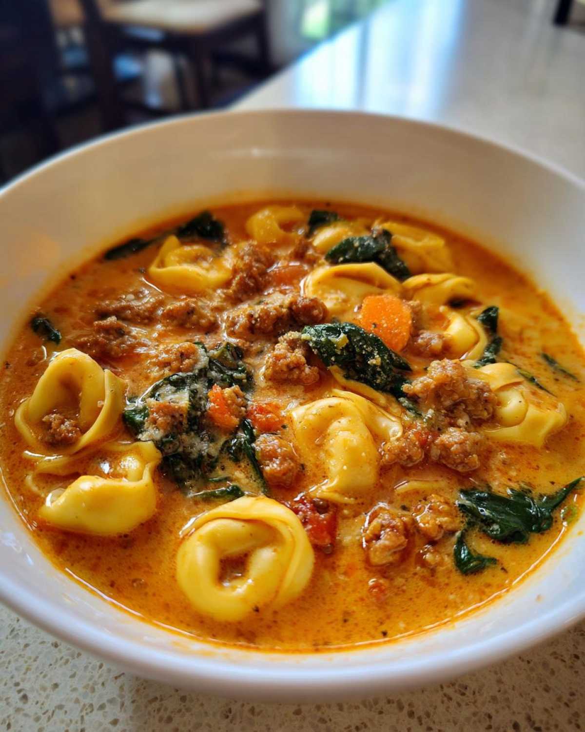 Close-up of a white bowl filled with creamy sausage tortellini soup, featuring tortellini, ground sausage, spinach, and carrots.