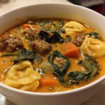 Close-up of a white bowl filled with creamy sausage tortellini soup, featuring sausage meatballs, spinach, and carrots.