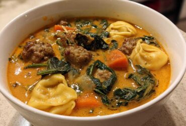 Close-up of a white bowl filled with creamy sausage tortellini soup, featuring sausage meatballs, spinach, and carrots.