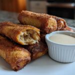 A pile of golden brown, crispy peach cobbler egg rolls served with a small white ramekin of creamy dipping sauce.
