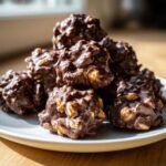 A pile of freshly made crockpot peanut clusters coated in rich chocolate, sitting on a white plate.