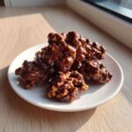A mound of glossy, freshly made crockpot peanut clusters piled on a small white plate near a window.