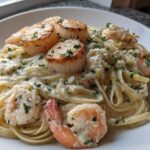 Close-up of creamy seafood pasta featuring fettuccine, seared scallops, and pink shrimp, garnished with parsley.