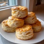 A stack of six golden-brown, flaky buttermilk biscuits topped with a light glaze, resting on a white plate.
