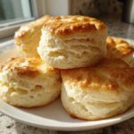 A stack of four golden-brown, flaky buttermilk biscuits resting on a white plate near a sunny window.