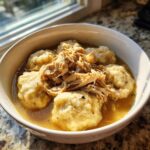 A white bowl filled with rich broth, fluffy dumplings, and shredded chicken, representing classic chicken and dumpling.