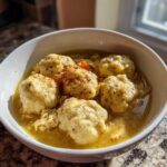 A white bowl filled with rich broth, shredded chicken, and five fluffy dumplings, seasoned with pepper, representing classic chicken and dumpling.