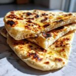 Close-up of stacked, fluffy homemade naan bread pieces brushed with melted butter or ghee, showing charred spots.
