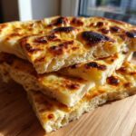 A stack of freshly baked, fluffy homemade naan bread pieces with charred, golden-brown spots, resting on a wooden board.