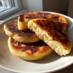 A stack of freshly cooked, golden-brown irish potato farls on a white plate, one cut open showing the fluffy interior.
