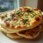 A stack of freshly baked, soft naan bread topped with melted butter and chopped green herbs.