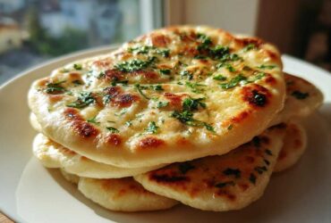 A stack of freshly baked, soft naan bread topped with melted butter and chopped green herbs.